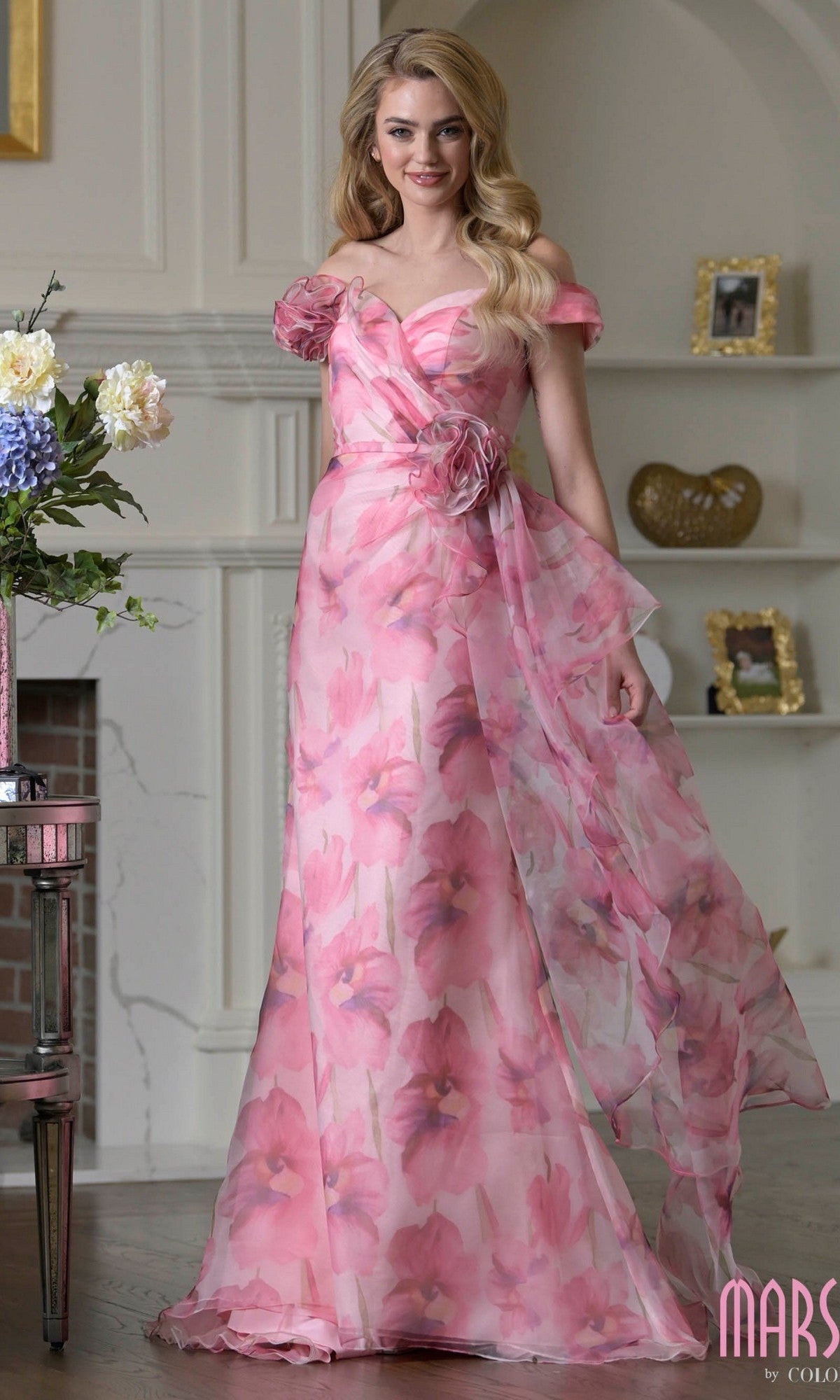 A woman indoors stands beside a floral vase and shelf, wearing the Long Formal Dress: Marsoni MV1394 in Pink Multi—an off-shoulder, pink floral formal dress with a flowing sheer overlay.